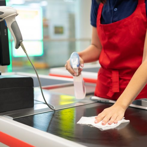 Unrecognizable cashier wearing red apron cleansing checkout surface with disinfectant in modern store