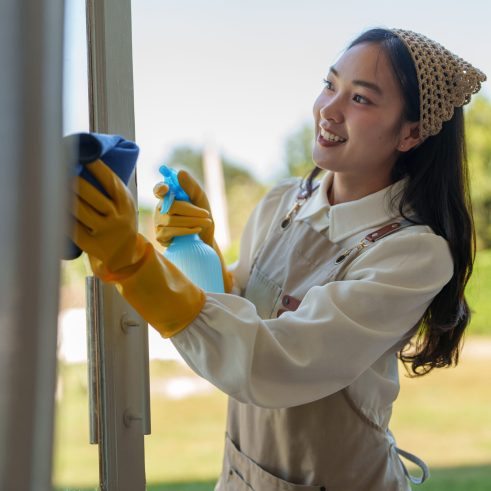 Smiling housekeeper wearing yellow gloves and apron cleaning window glass with spray bottle and rag, enjoying her work in a bright and airy home environment